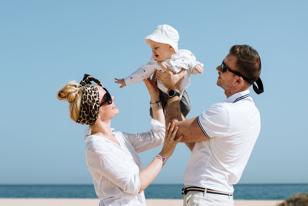 Happy family with parents lifting smiling baby at sunny beach, wearing white outfits against blue sky.