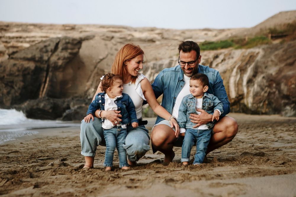 Family of four posing on sandy beach, parents with two toddlers in matching denim outfits, rocky cliffs in background.