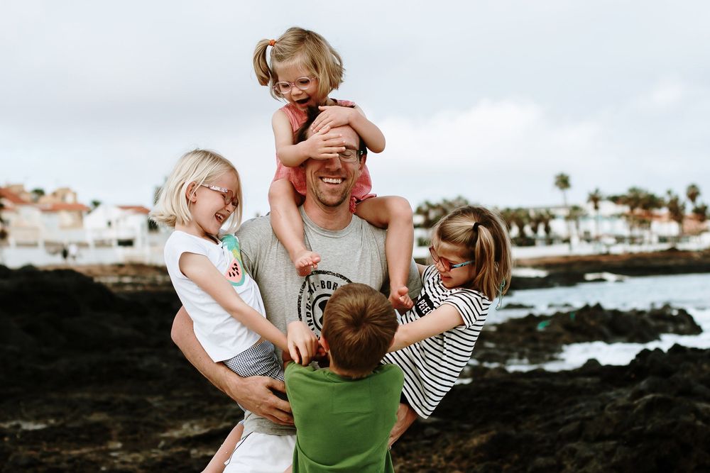 A happy father plays with four young children near a rocky seaside, one child sitting on his shoulders covering his eyes.