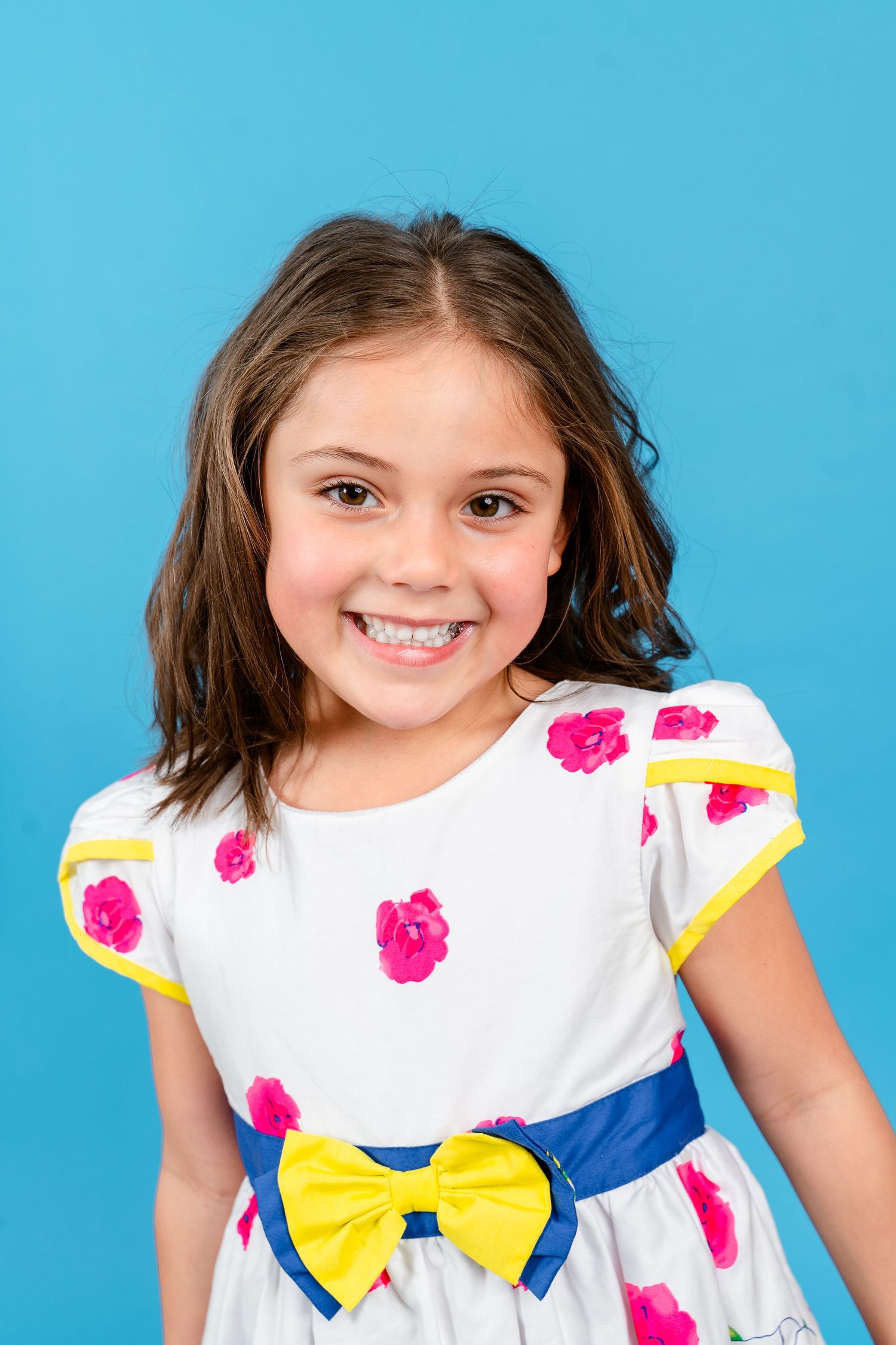 studio portrait of a smiling young brunette girl, wearing a white dress, against a bright blue background