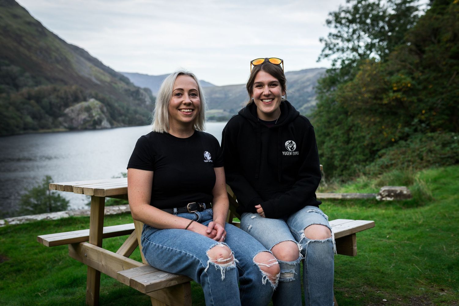 Two smiling women in black tops sit on a wooden bench beside a scenic lake surrounded by green hills.