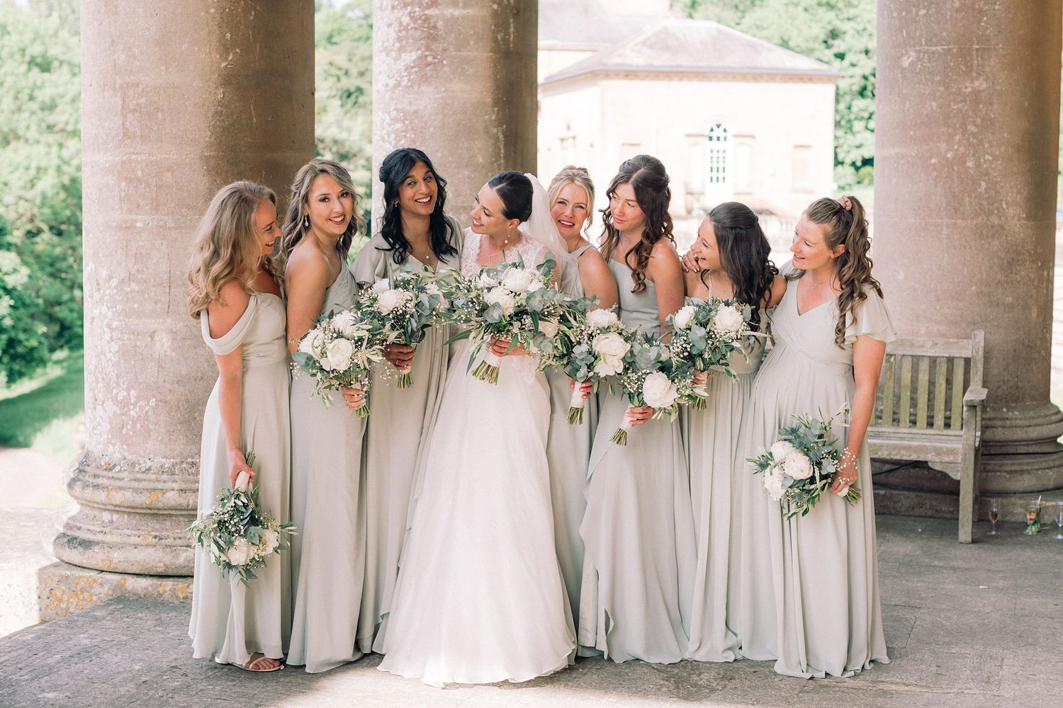 Bride in white gown with six bridesmaids in sage green dresses holding white floral bouquets at elegant venue.