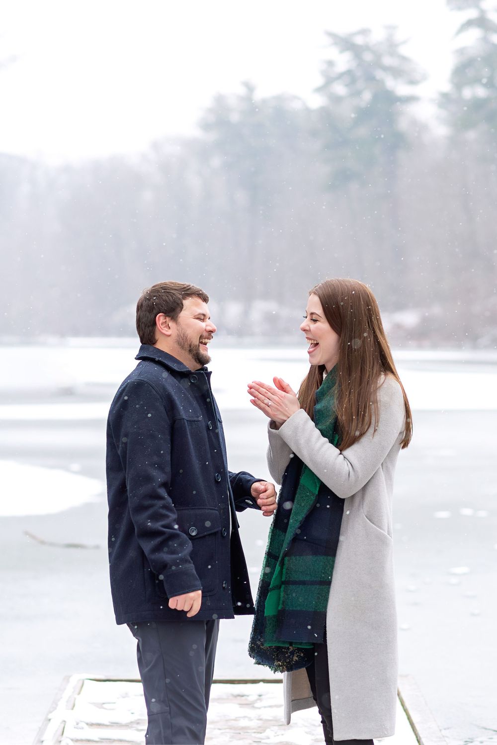 Couple laughing together on snowy dock by frozen lake in winter, woman wearing gray coat and green plaid scarf.