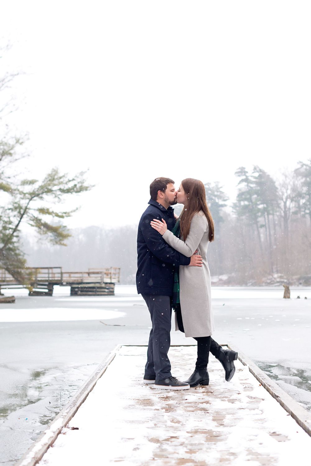 Couple kissing on snowy dock by frozen lake in winter, wearing elegant coats during snowfall.