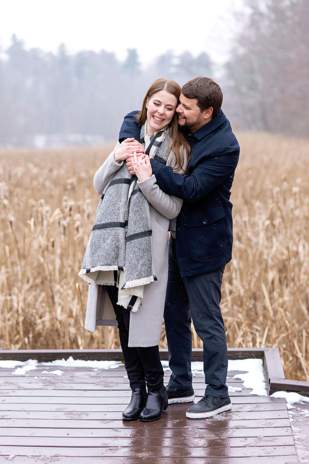 Couple embracing on a snowy dock in winter, woman holding a red cup while man hugs her from behind near a reed field.