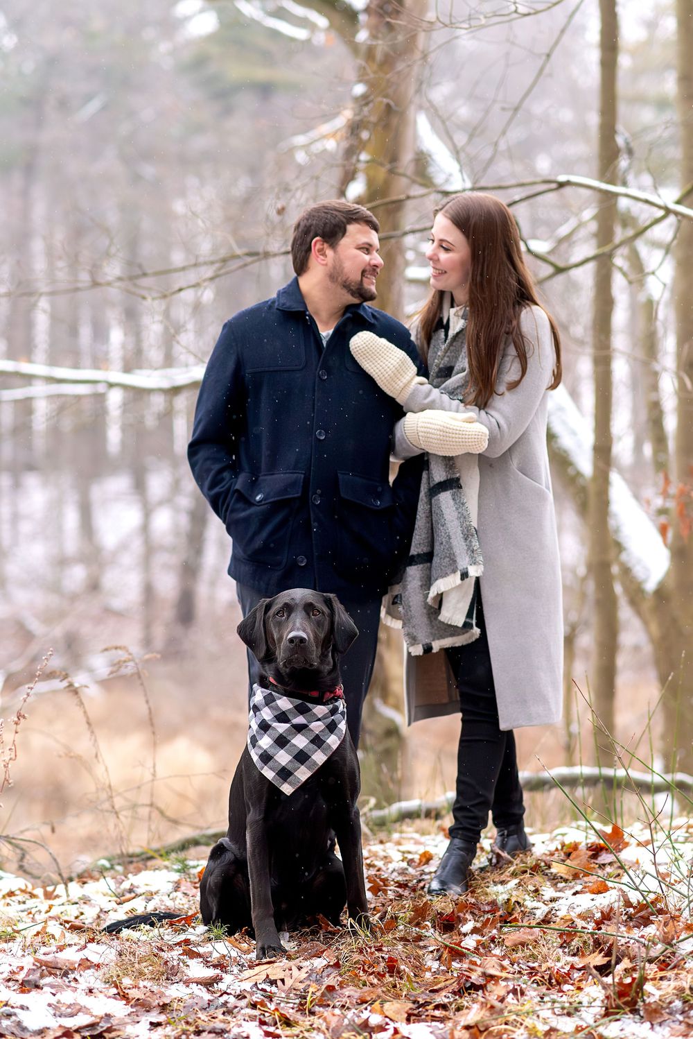 Couple smiling at each other in snowy woods with their black Labrador dog sitting in front.