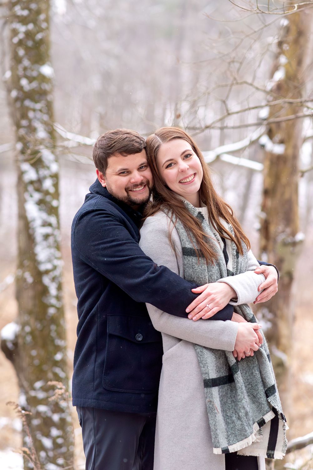 Couple embracing in snowy winter forest, man hugging woman from behind, both smiling at camera.