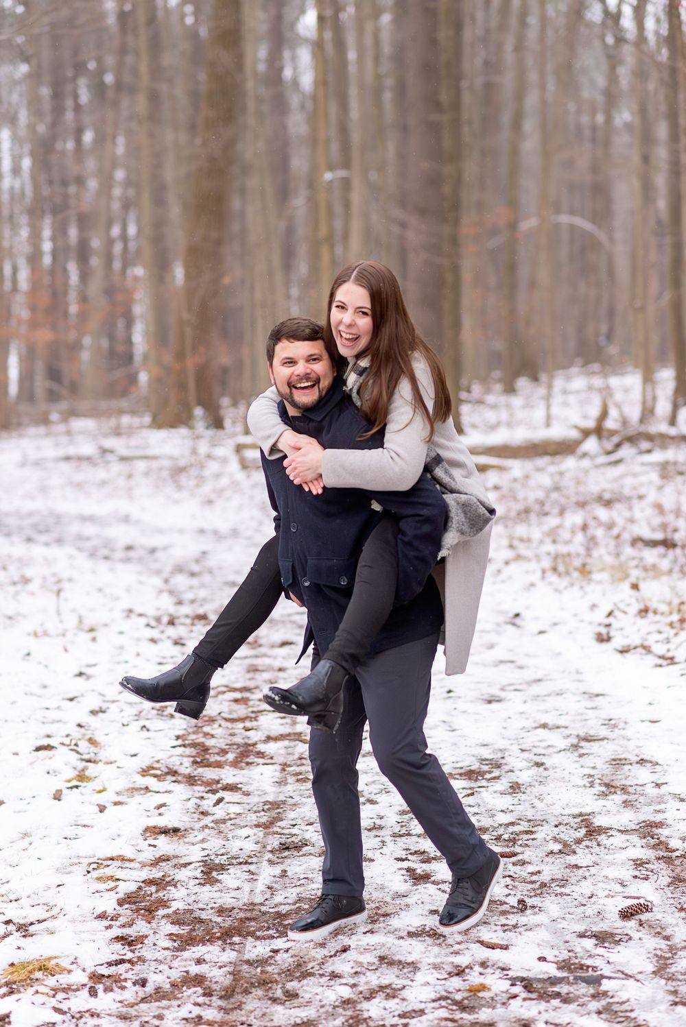 Laughing man giving woman a piggyback ride along a snow-covered forest path in winter.