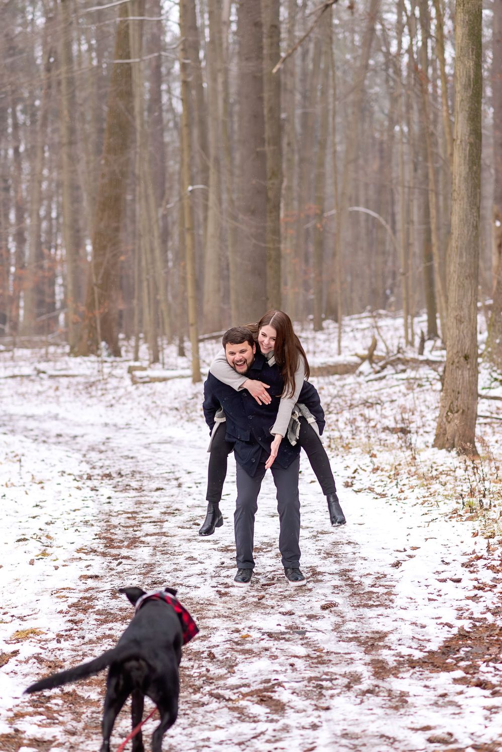 Man giving woman a piggyback ride in snowy woods with black dog nearby.