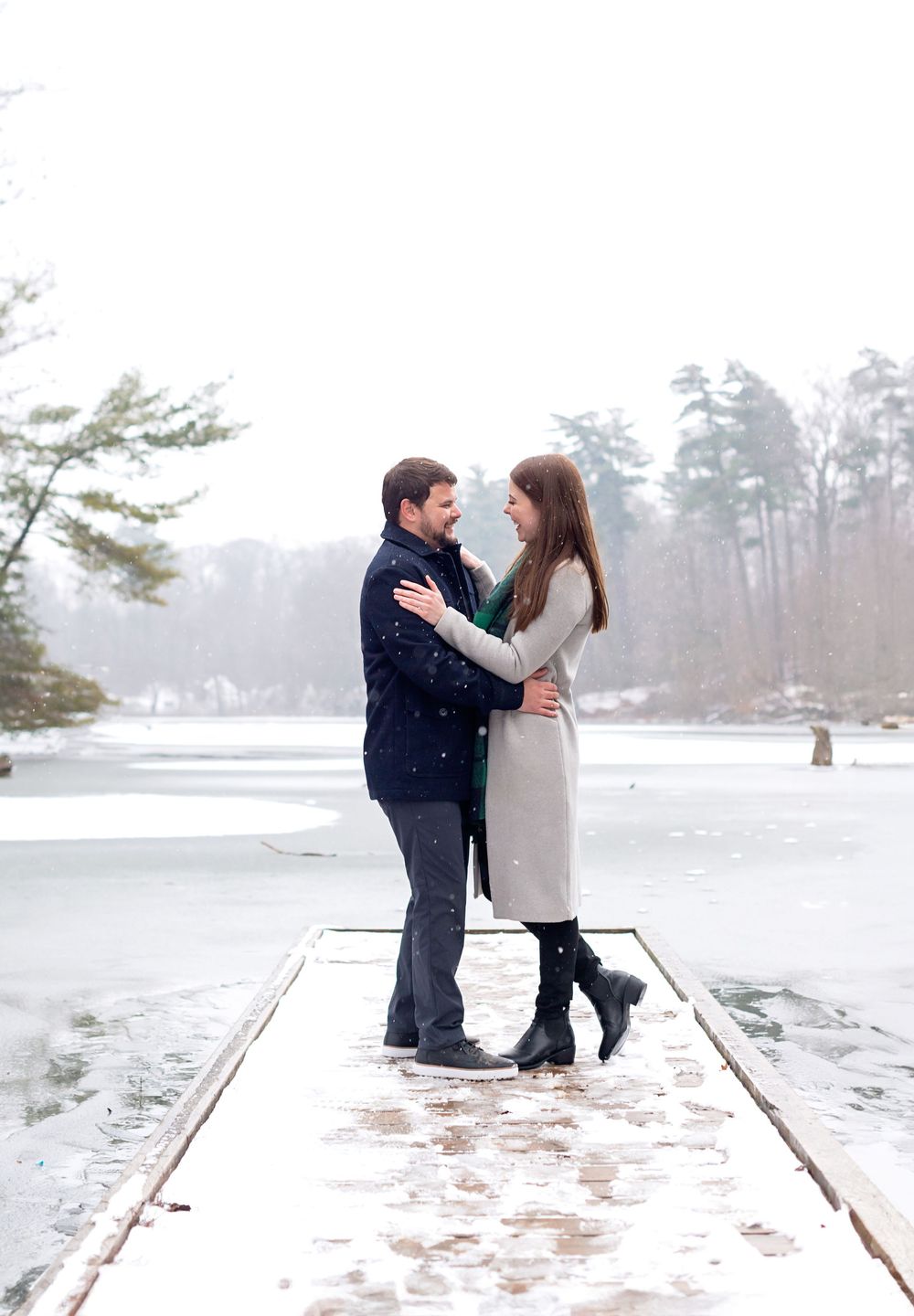 Couple embracing on a snowy dock by a frozen lake in winter, wearing coats and boots.