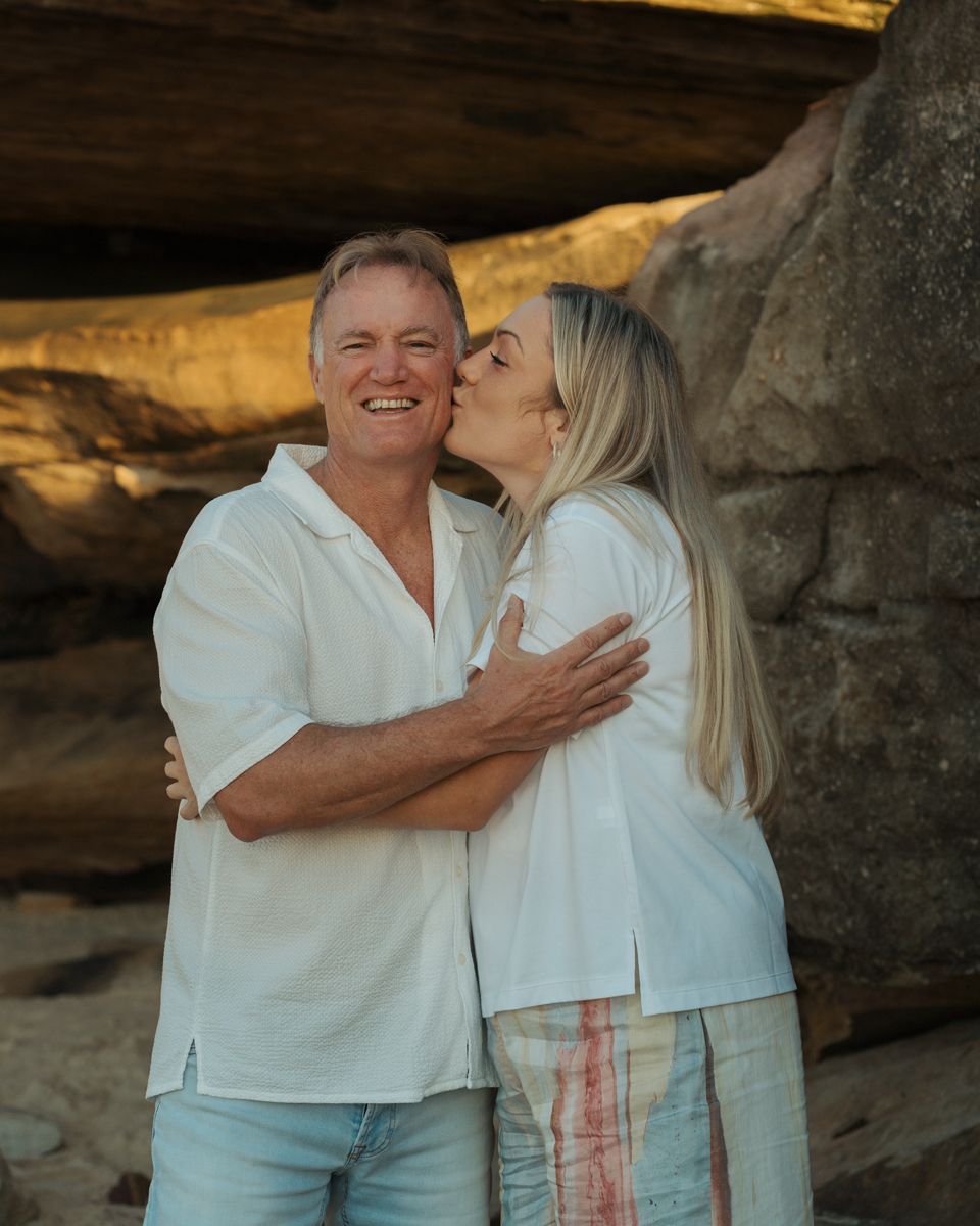 Father smiling while adult daughter kisses him on cheek near rocky cliffs at Bronte Beach family photoshoot