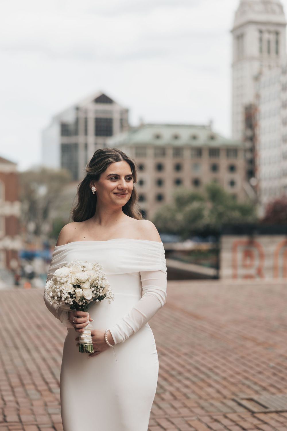 Bride in off-shoulder white gown holds bouquet on brick plaza with city buildings backdrop.