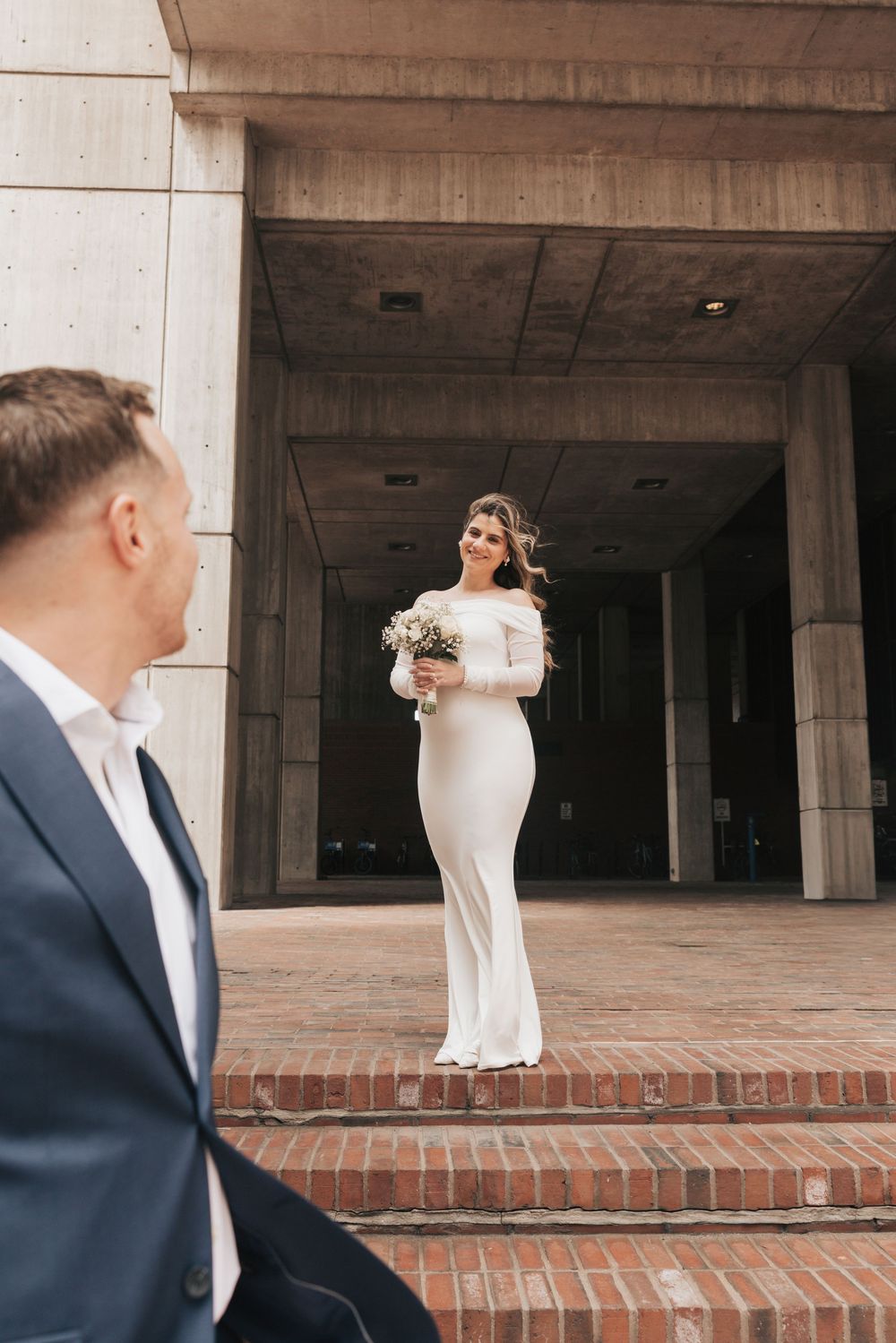 Bride in white off-shoulder gown holding bouquet stands on brick steps while groom in navy suit looks on.