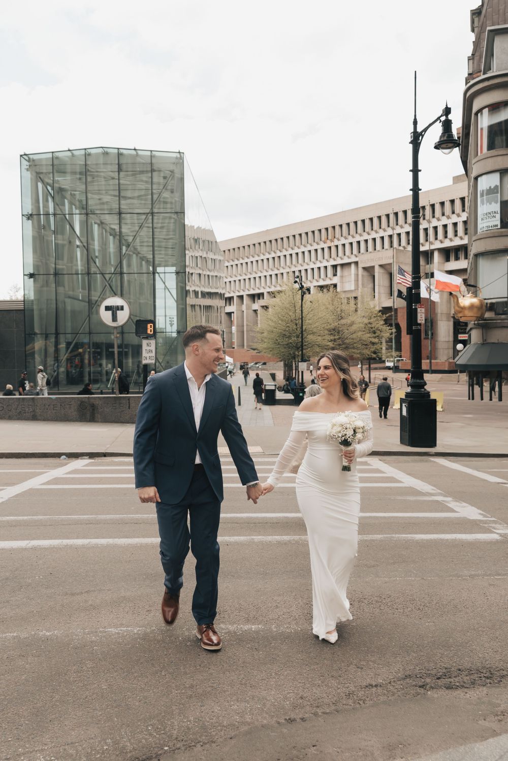 Bride and groom hold hands crossing a city street in Boston, bride in white off-shoulder gown holding bouquet.