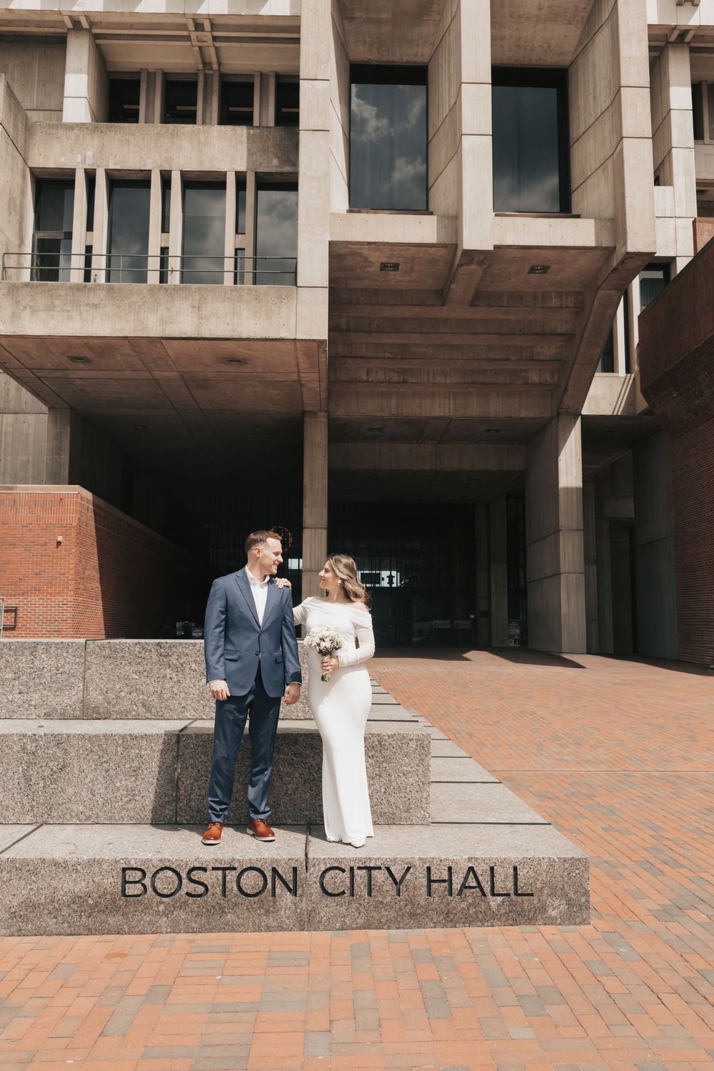 Bride and groom pose on steps of Boston City Hall, she in white gown holding bouquet, he in blue suit.