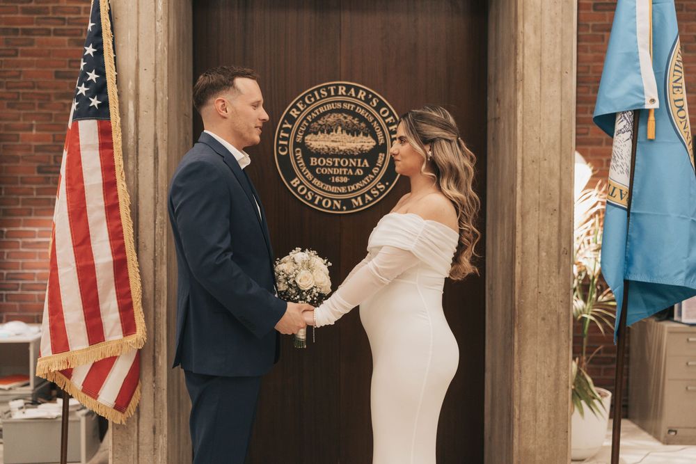 Couple exchanging vows at Boston City Hall Registry, bride in white off-shoulder gown, groom in navy suit.