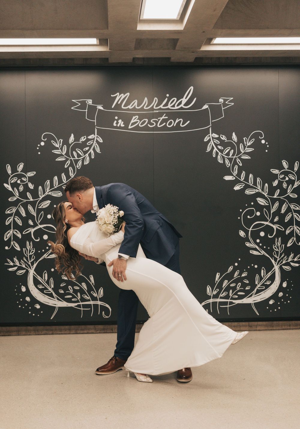 Newlyweds share a romantic dip kiss in front of a Married in Boston chalkboard mural backdrop.