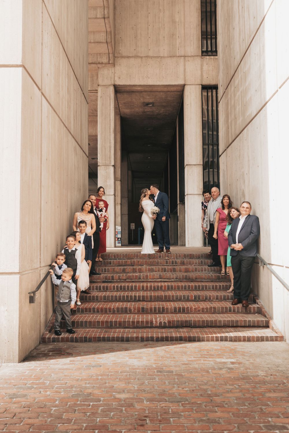 Wedding party poses on brick steps of a grand building as bride and groom share a kiss in the doorway.