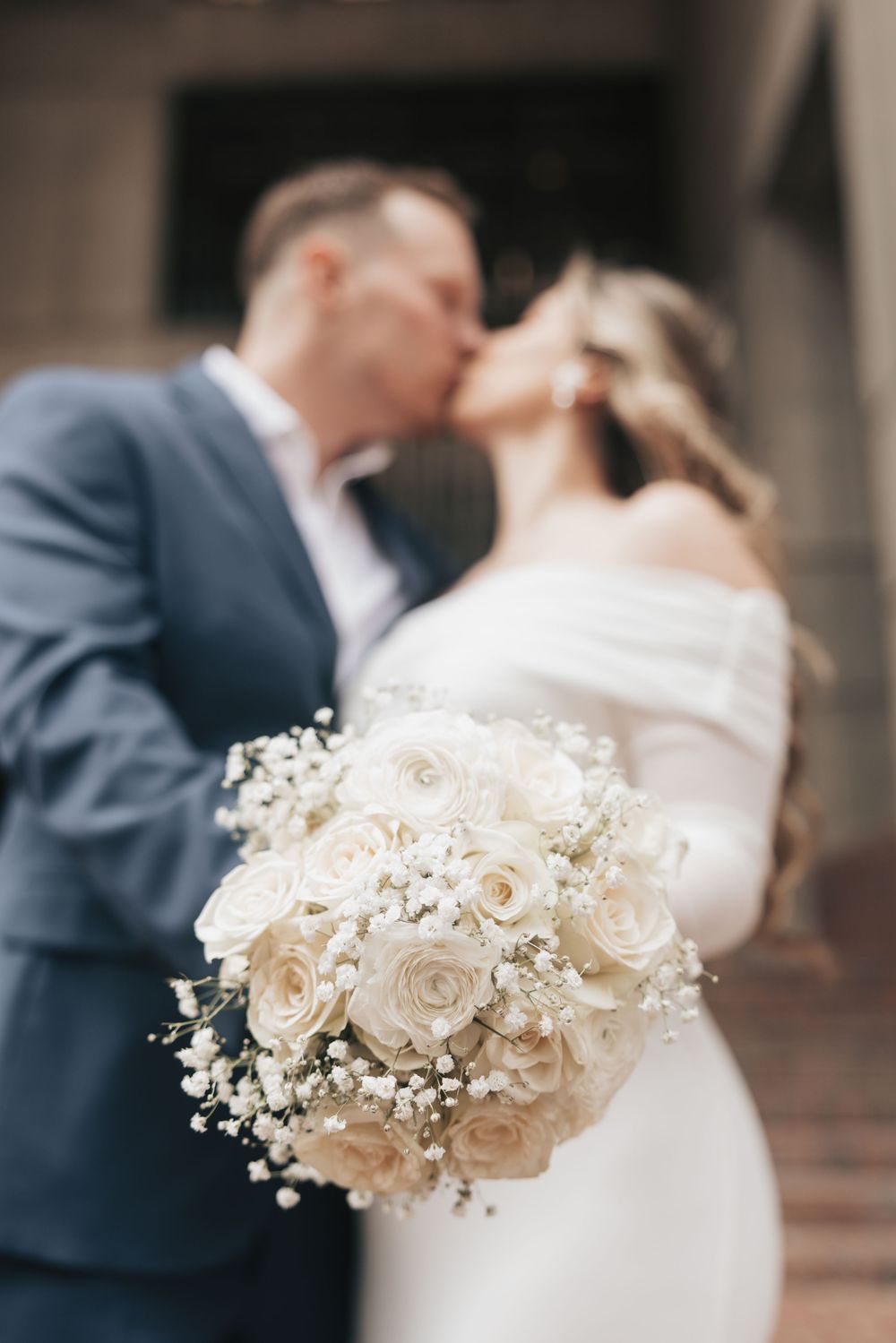 Newlyweds share a kiss behind a stunning white rose and baby's breath wedding bouquet in soft focus.