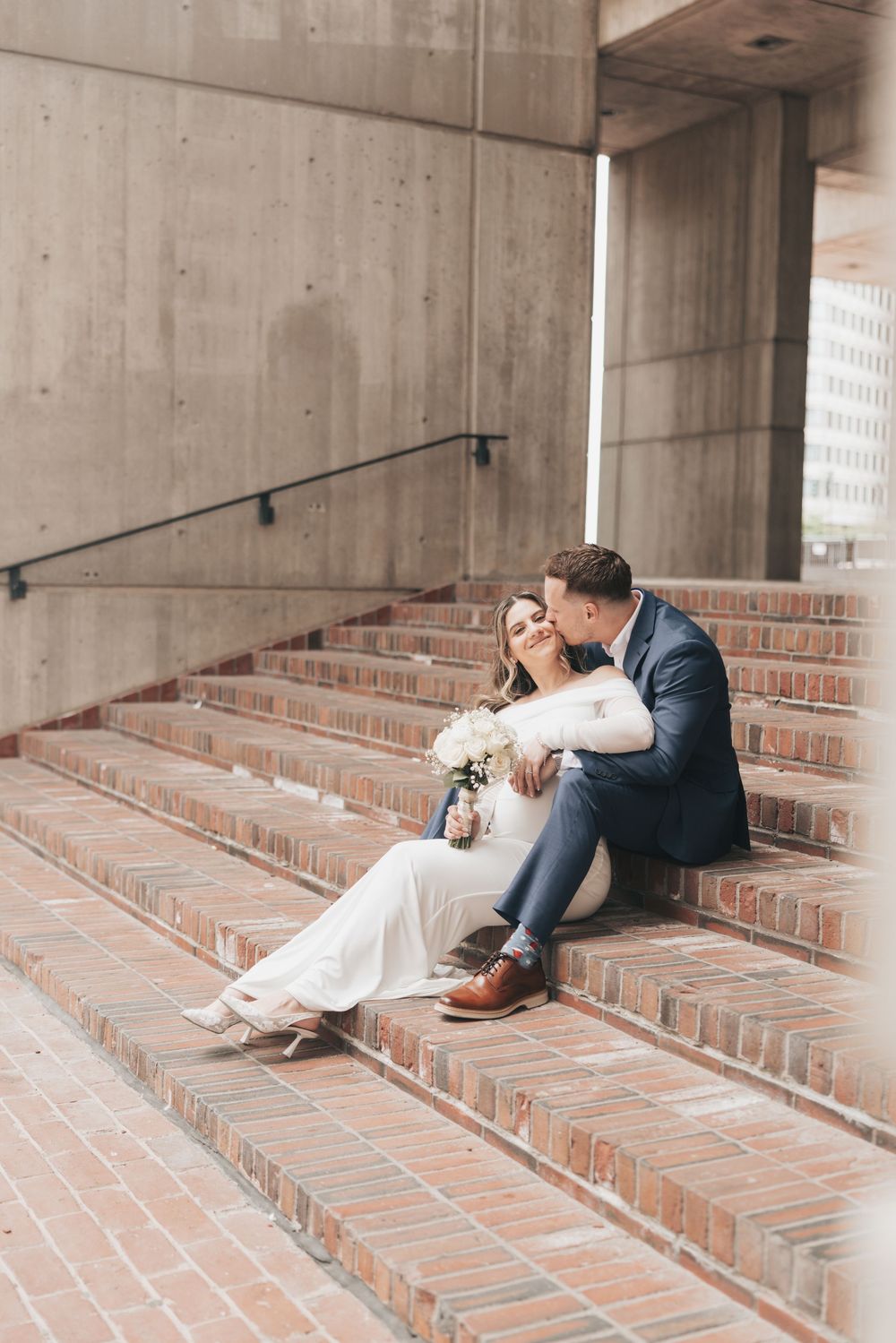 Bride and groom sit closely on brick steps, she holds white bouquet, he kisses her cheek, concrete architecture backdrop.