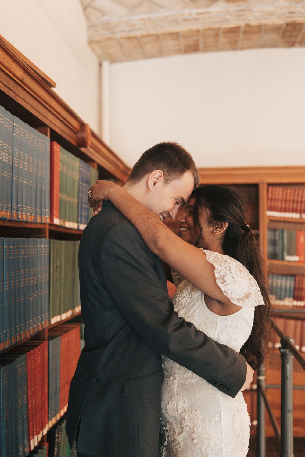 Bride and groom embracing romantically in a library, surrounded by colorful bookshelves, sharing a joyful wedding moment.
