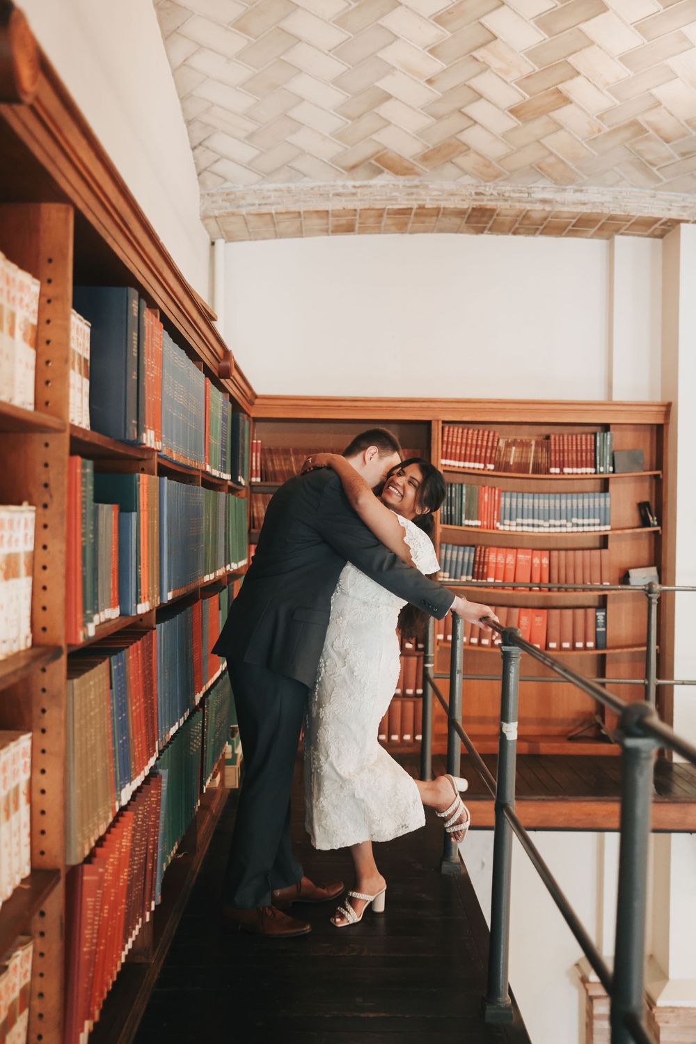 Couple embracing romantically on library stairs surrounded by wooden bookshelves filled with colorful books.