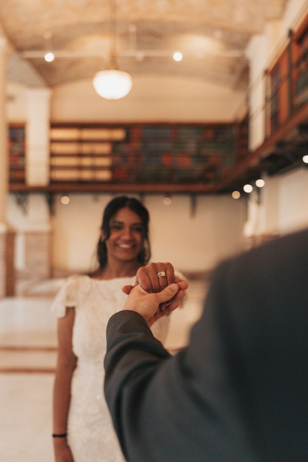 Smiling bride in white dress holds hands with groom in suit inside elegant venue with warm lighting.