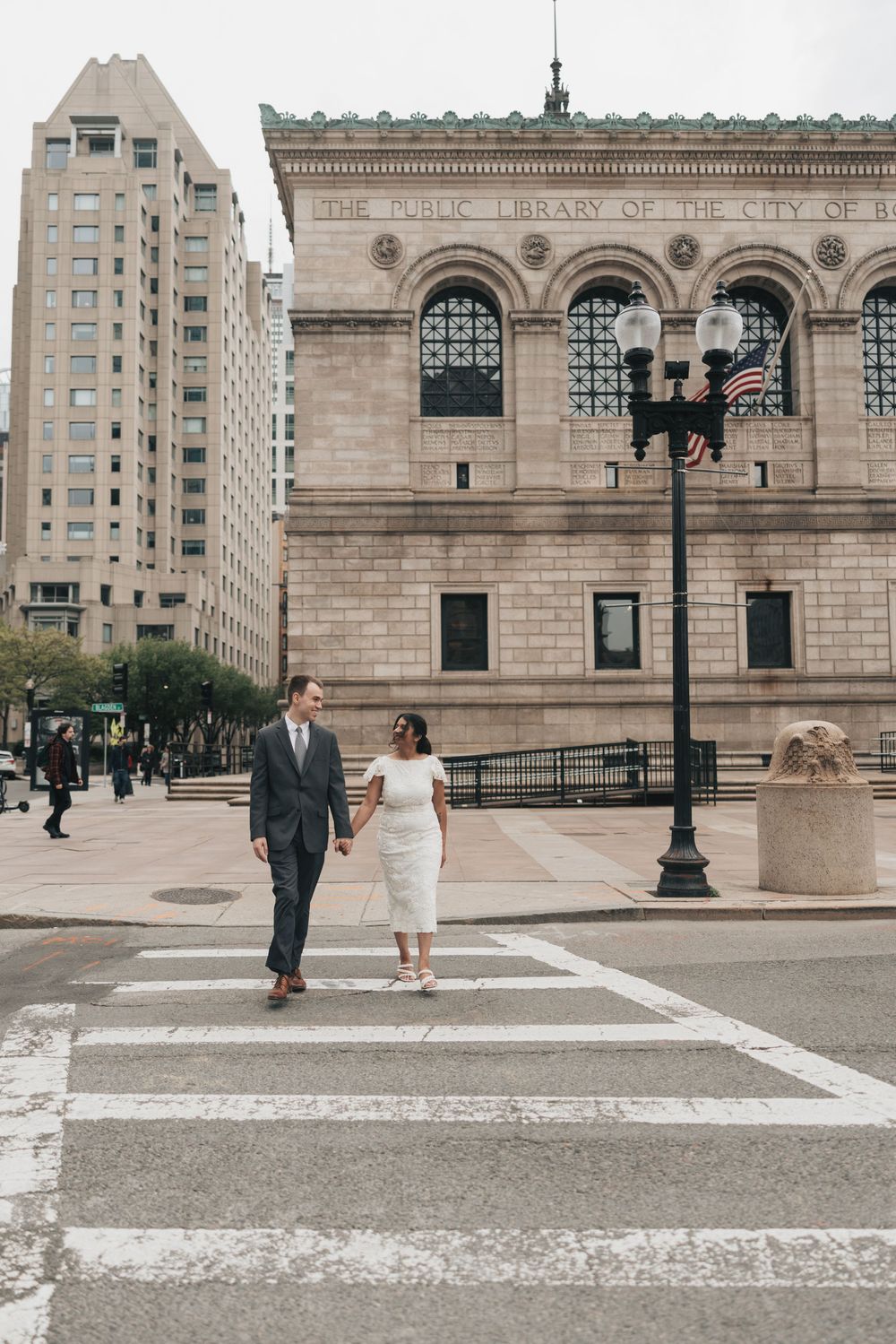 Couple in wedding attire holds hands crossing a zebra crossing in front of a grand city library building.