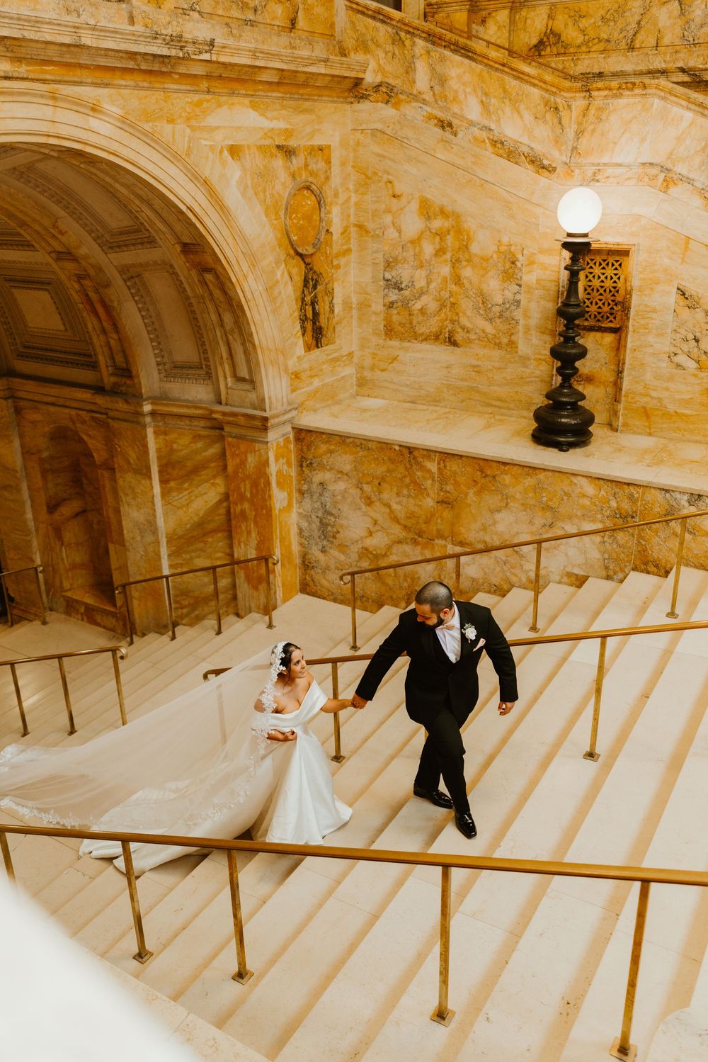 Bride and groom walking up grand marble staircase with gold railings in elegant wedding venue.
