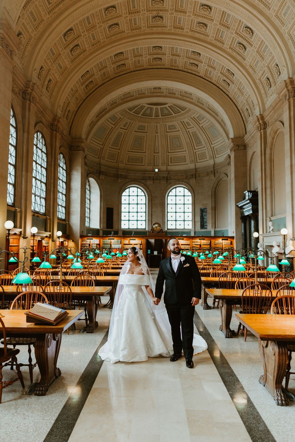 Bride and groom hold hands in ornate Boston Public Library reading room with arched ceiling and green lamp-lit tables.