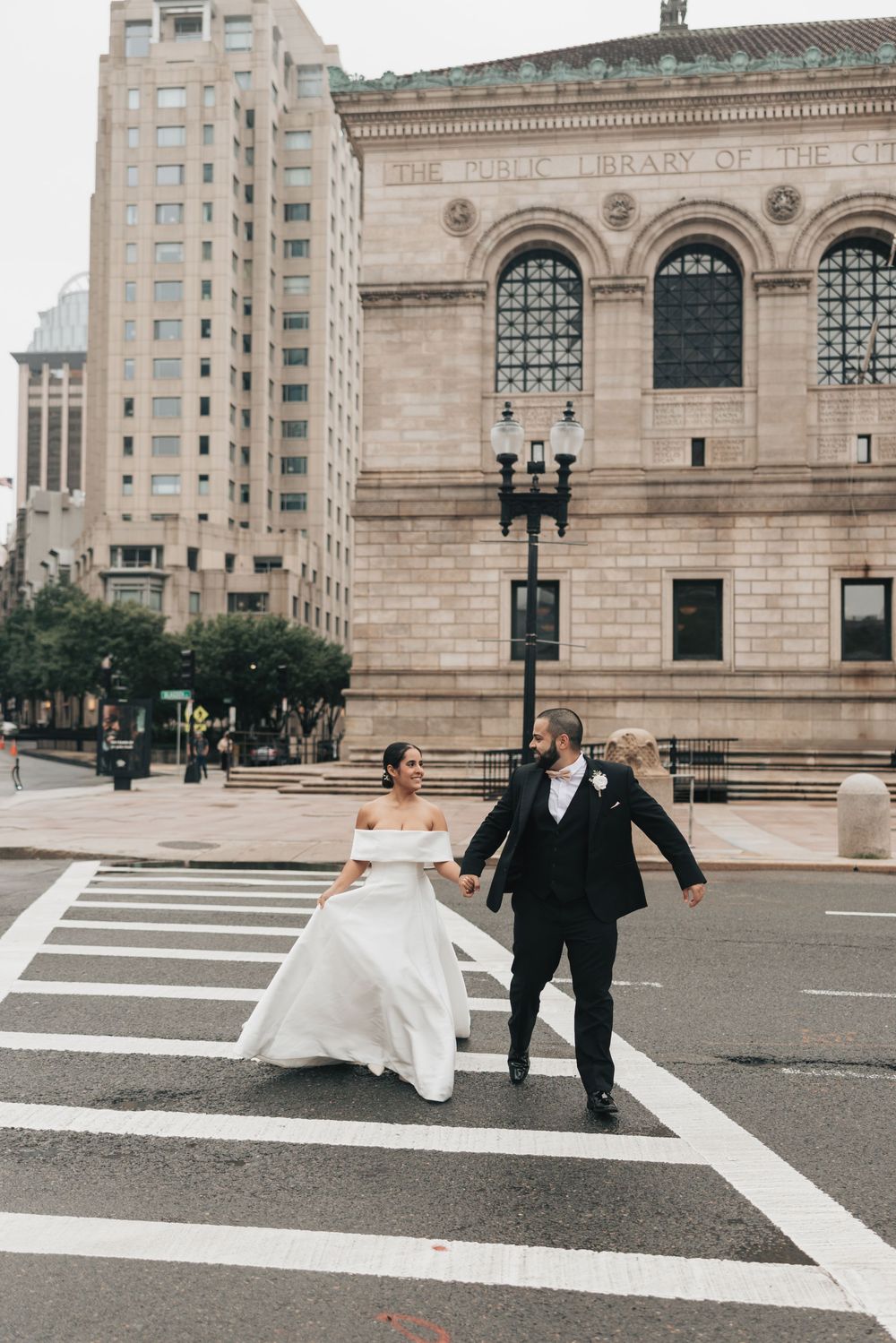 Bride and groom crossing a city street in wedding attire near a classic stone public library building.