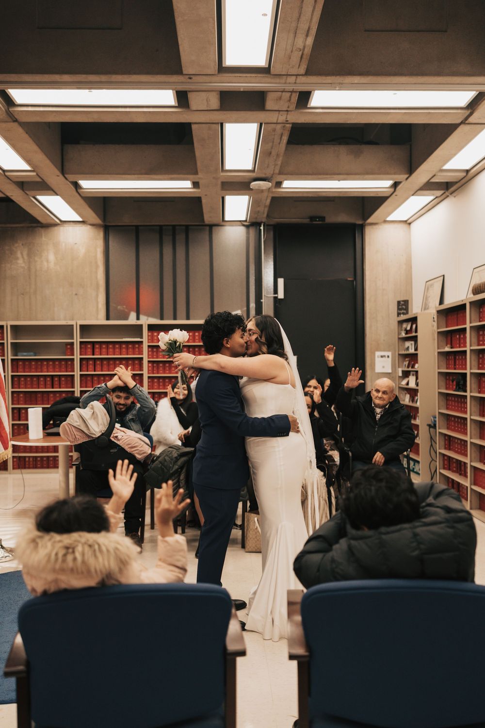 Couple shares first kiss at courthouse wedding ceremony as guests cheer in a government office setting.