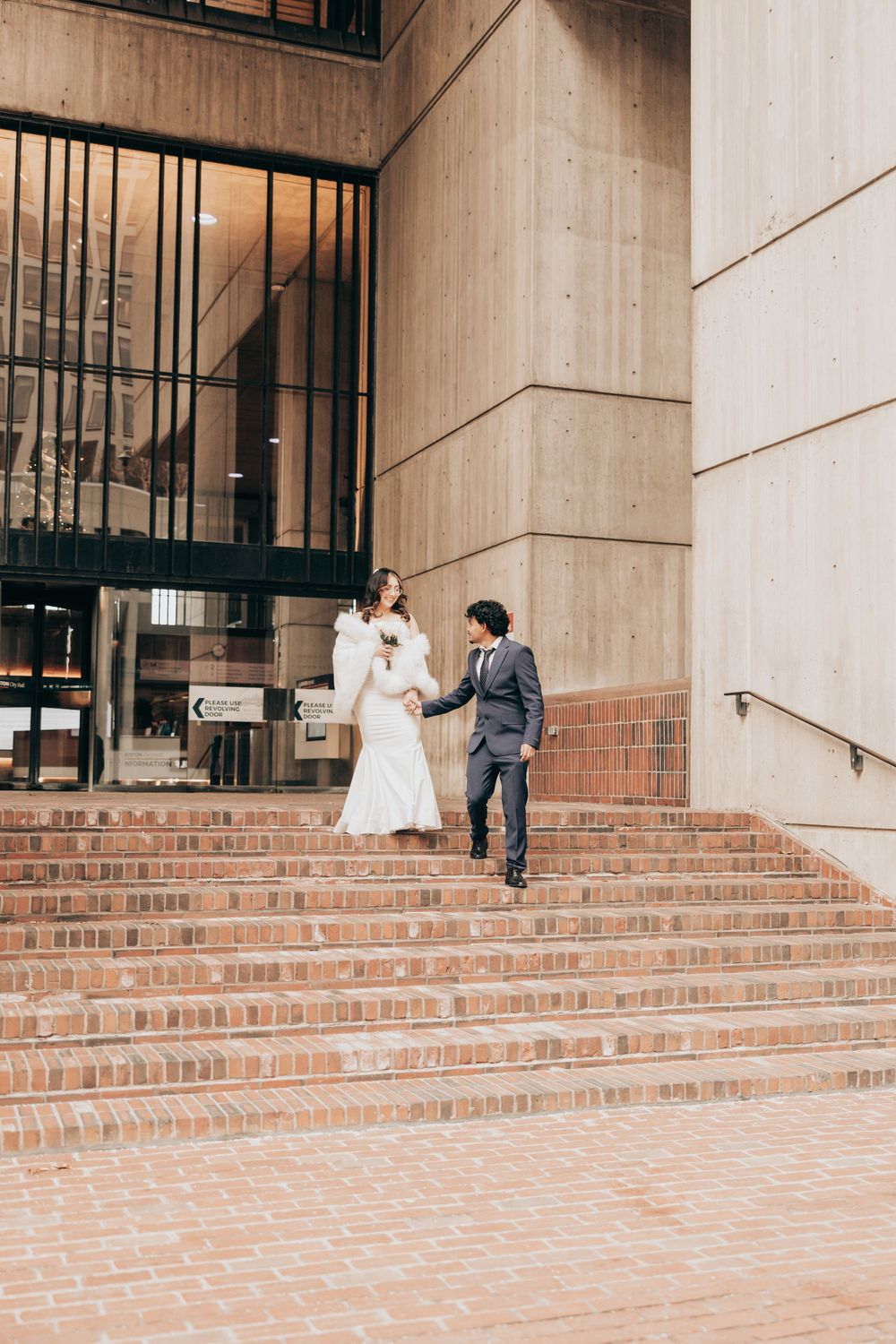 Bride and groom holding hands walking down brick steps outside a modern building on their wedding day.