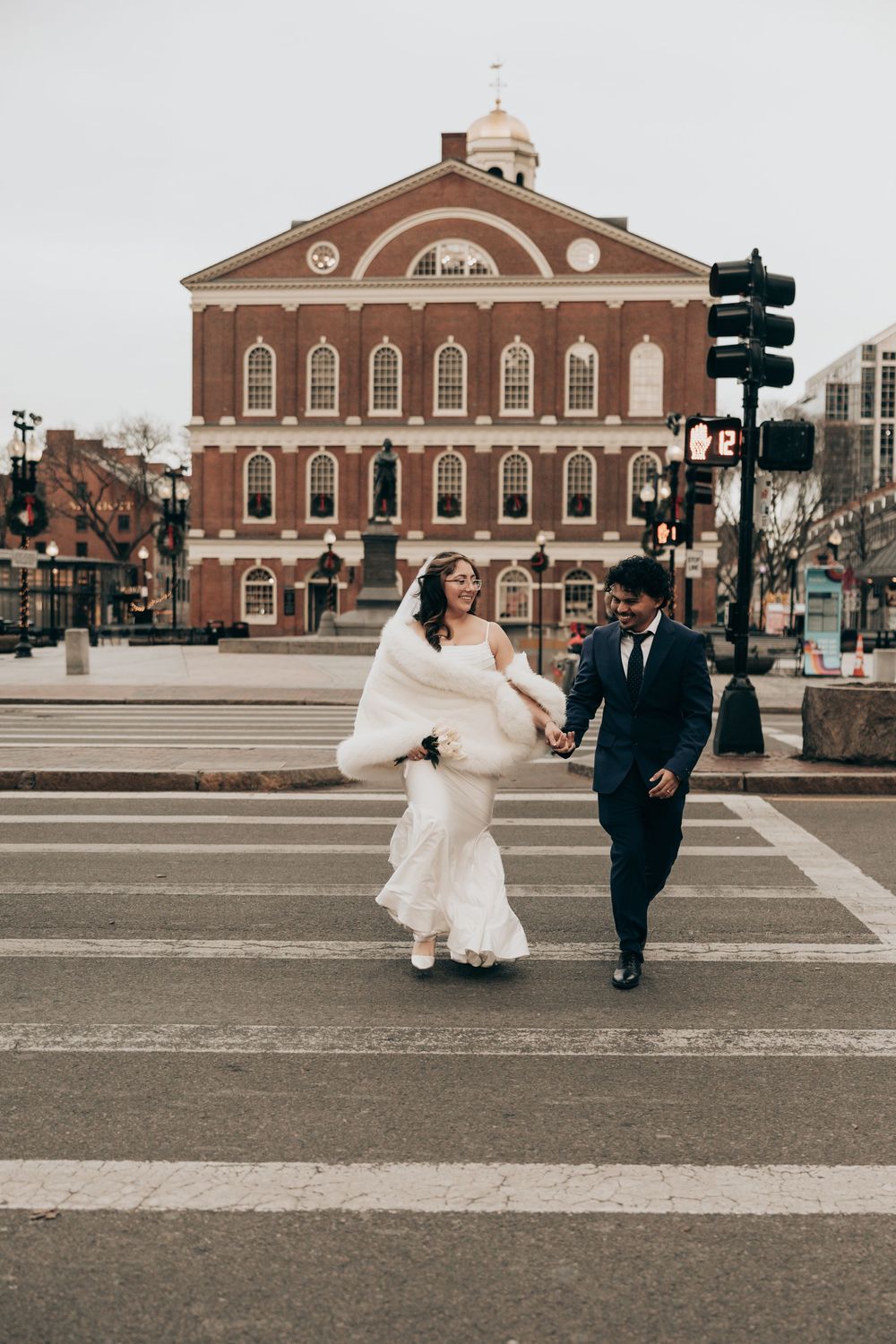 Bride and groom crossing street in front of Faneuil Hall, Boston, bride in white gown with fur wrap, groom in navy suit.