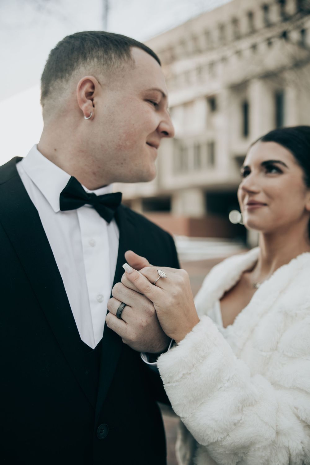 Bride and groom holding hands outdoors, wearing wedding rings, tuxedo and white fur wrap.