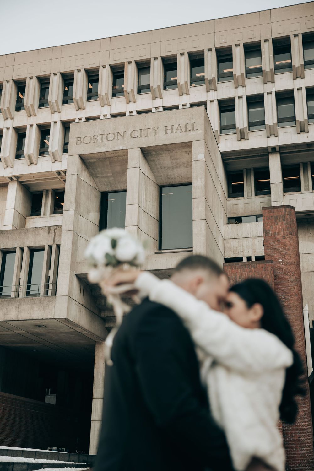 Couple kissing in front of Boston City Hall with a white wedding bouquet in the foreground.