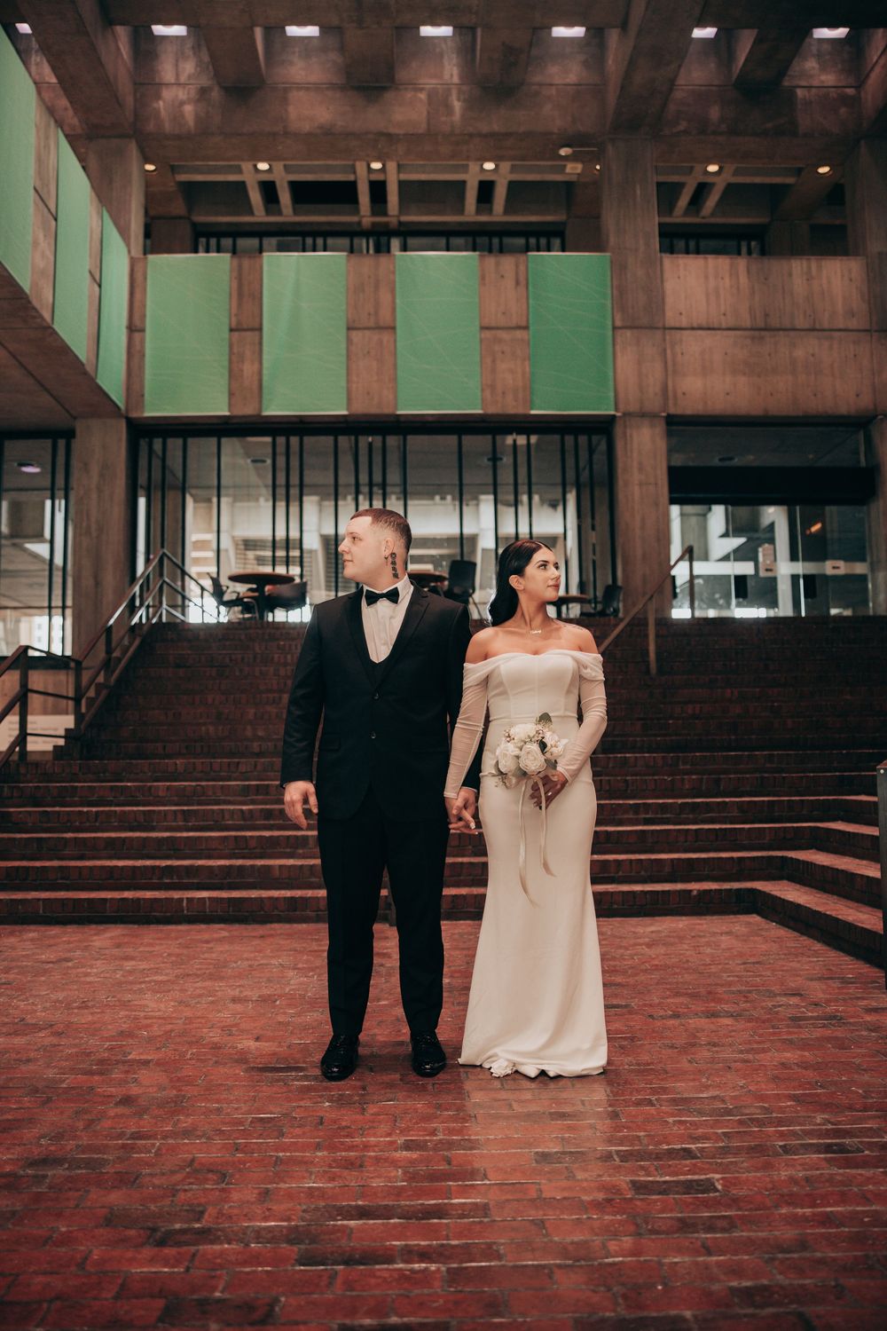 Bride and groom holding hands on brick steps outside a modern concrete building on their wedding day.
