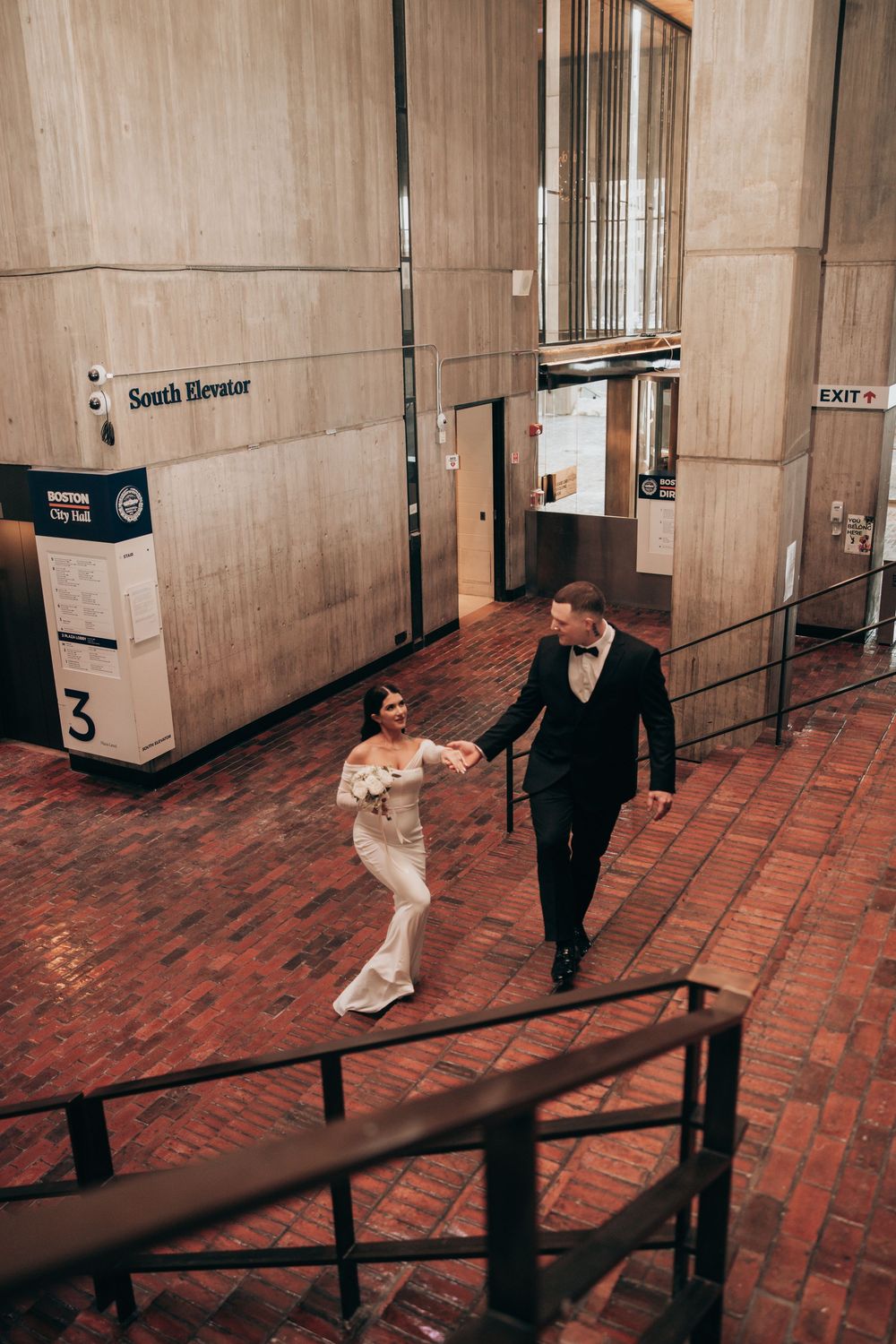 Bride and groom holding hands walking through Boston City Hall's brick interior near the South Elevator on their wedding day.