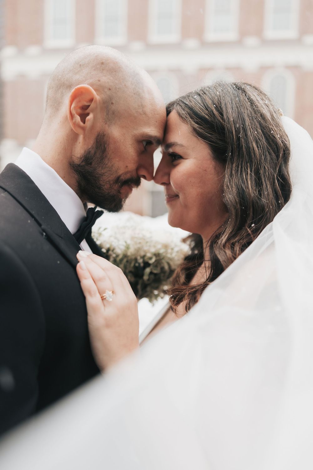 A bride and groom share an intimate forehead touch moment, with her veil flowing around them as she holds a white bouquet.