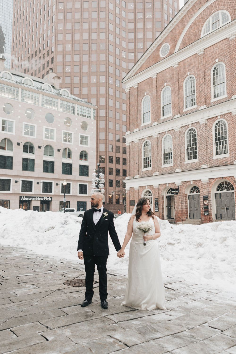 Bride and groom hold hands in snowy Boston plaza near Faneuil Hall, she in white gown, he in black tuxedo.