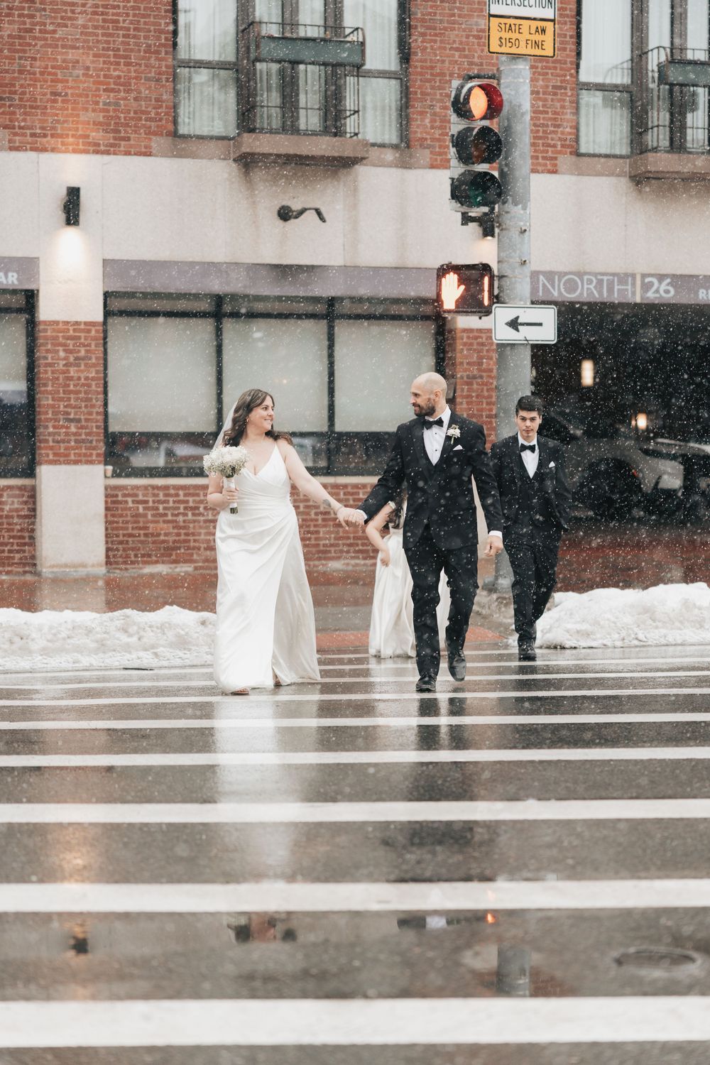 Bride and groom holding hands crossing a snowy urban street in winter, with a third person following behind.