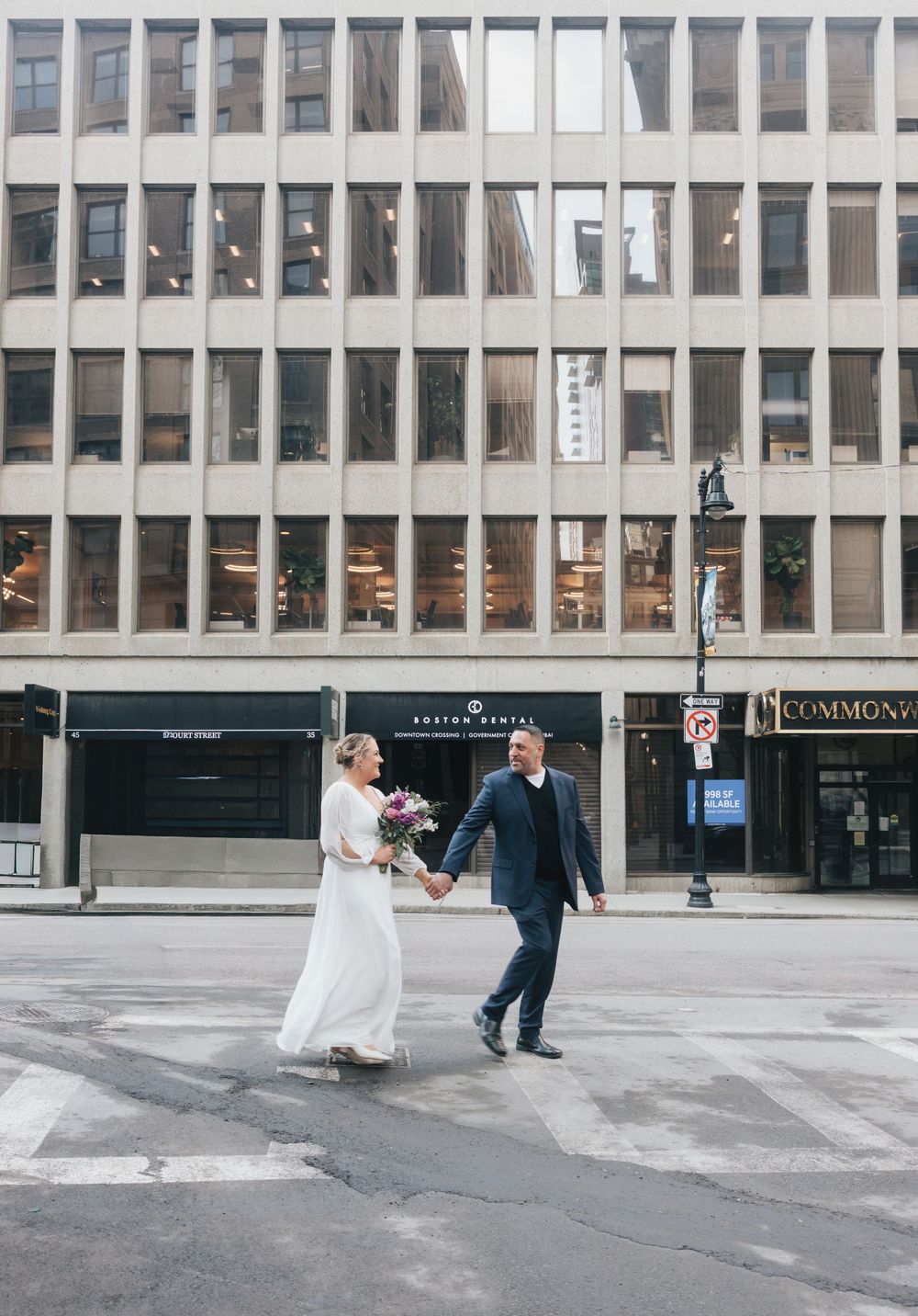 Bride and groom holding hands crossing a city street in front of a modern office building on their wedding day.