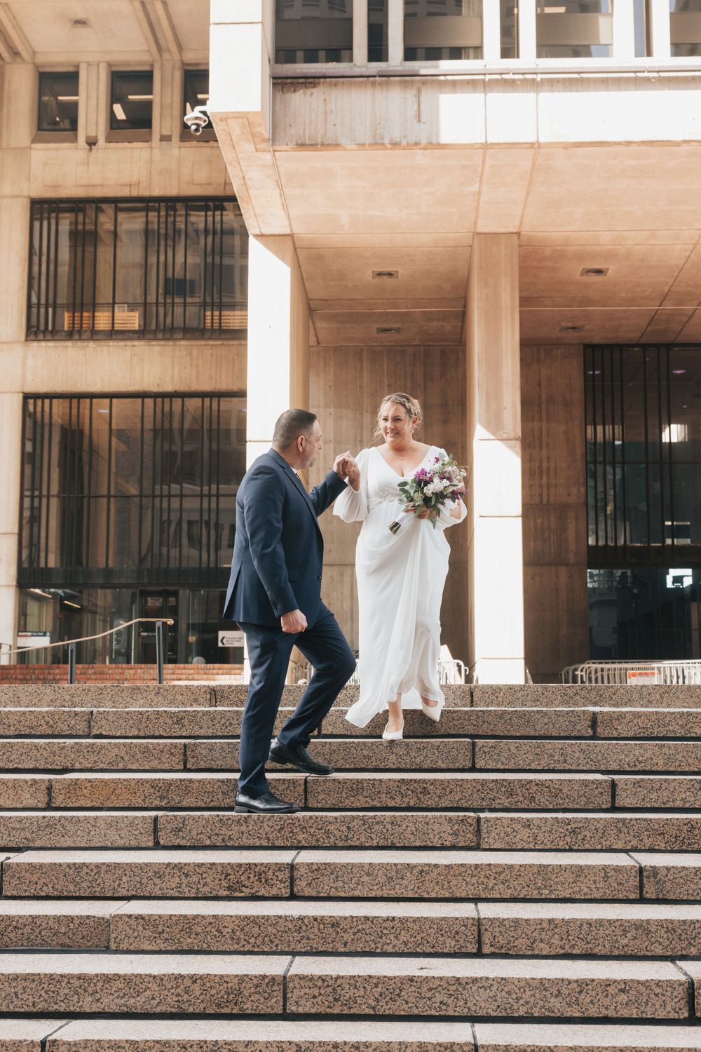 Bride and groom hold hands on stone steps outside a modern building, bride in white gown holding bouquet.
