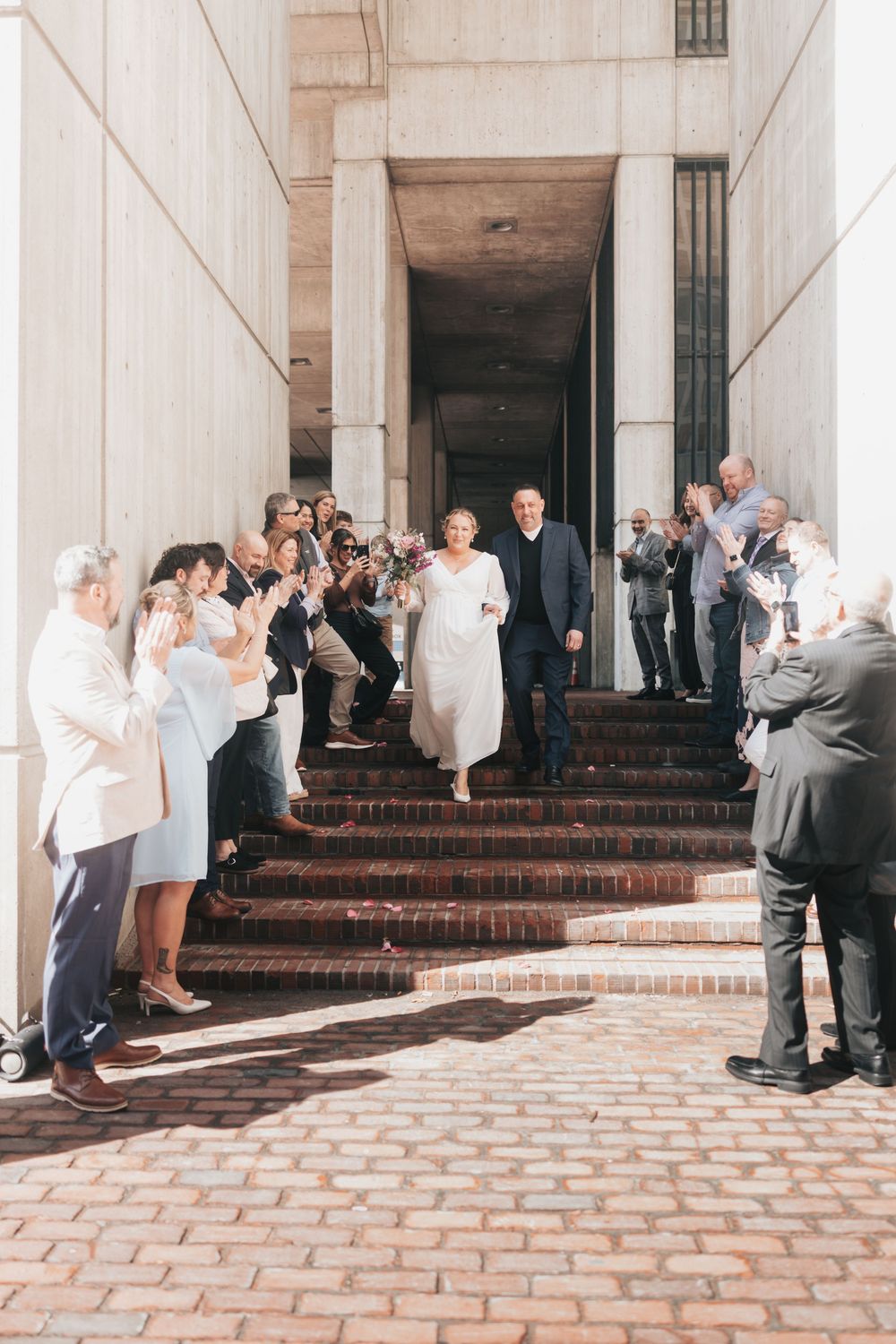 Newlywed couple descends brick steps while wedding guests applaud and celebrate outside a grand stone building.