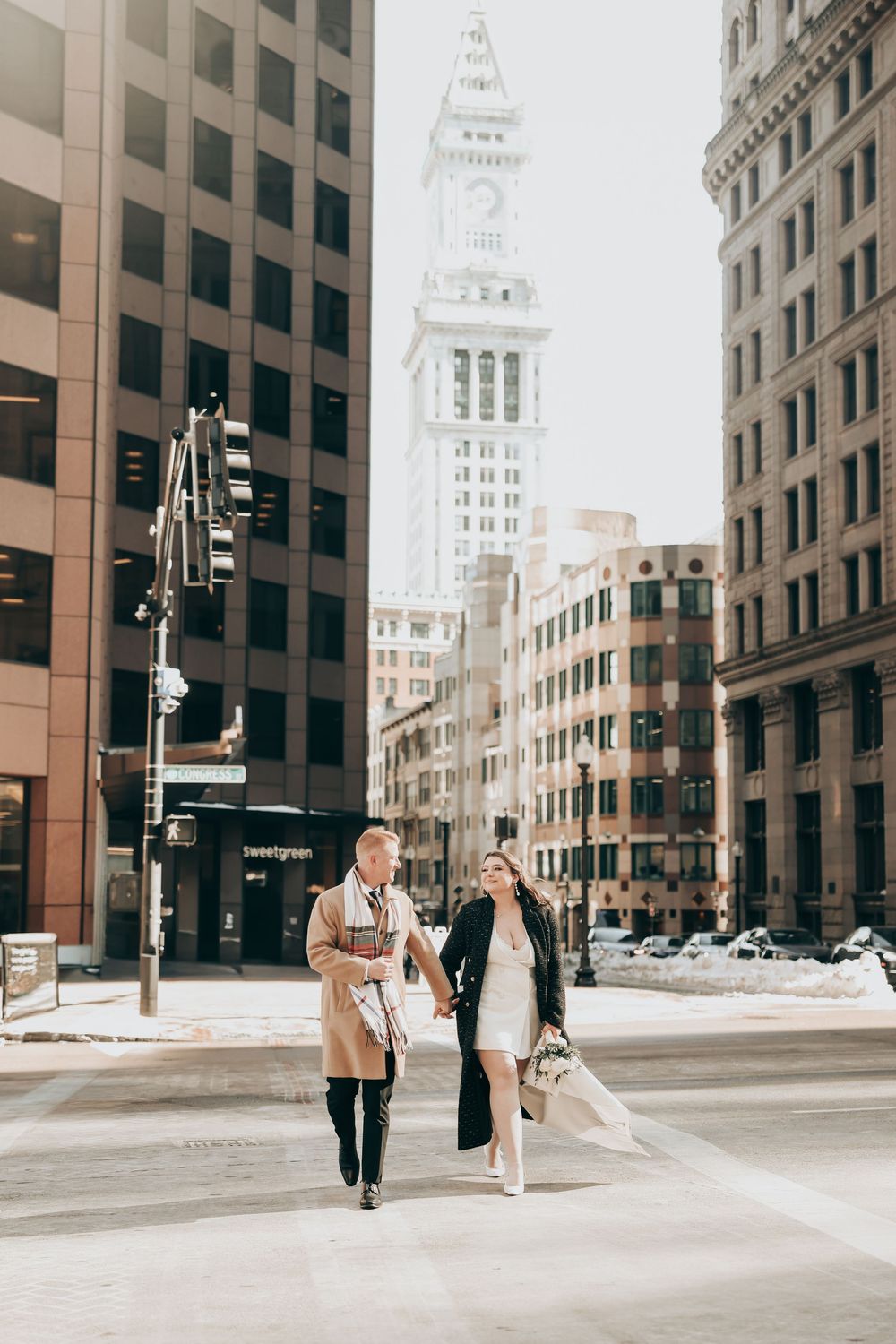 Couple walks hand-in-hand through snowy Boston streets with the Custom House Tower visible in the background.