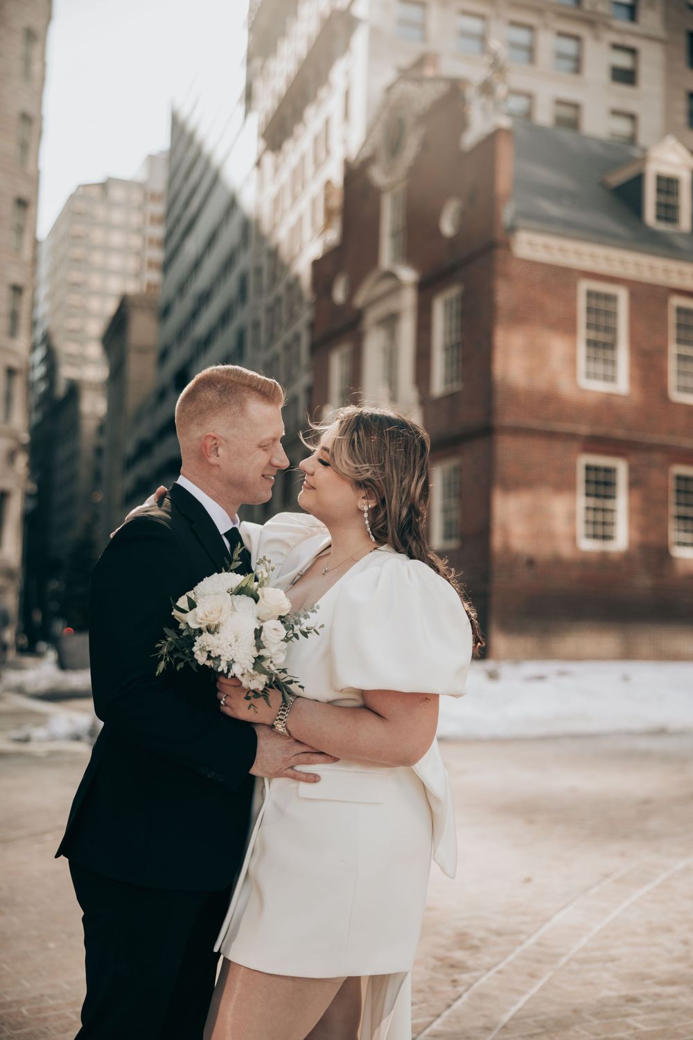 Bride and groom embrace in front of historic brick building in Boston, bride holding white bouquet in short wedding dress.