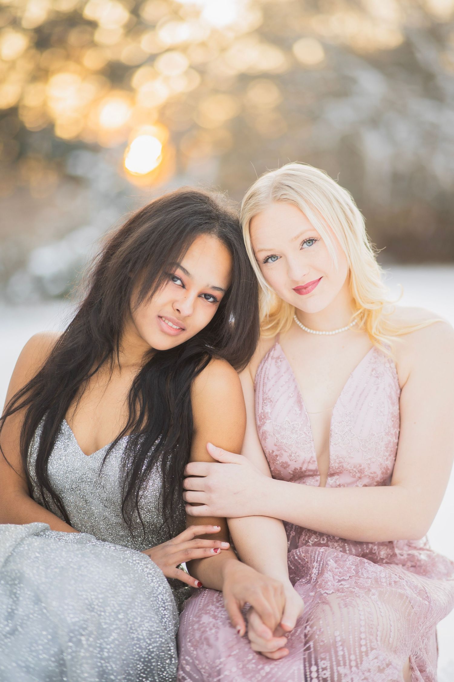 Two young women in elegant gowns pose together in a snowy winter landscape with golden sunset bokeh.