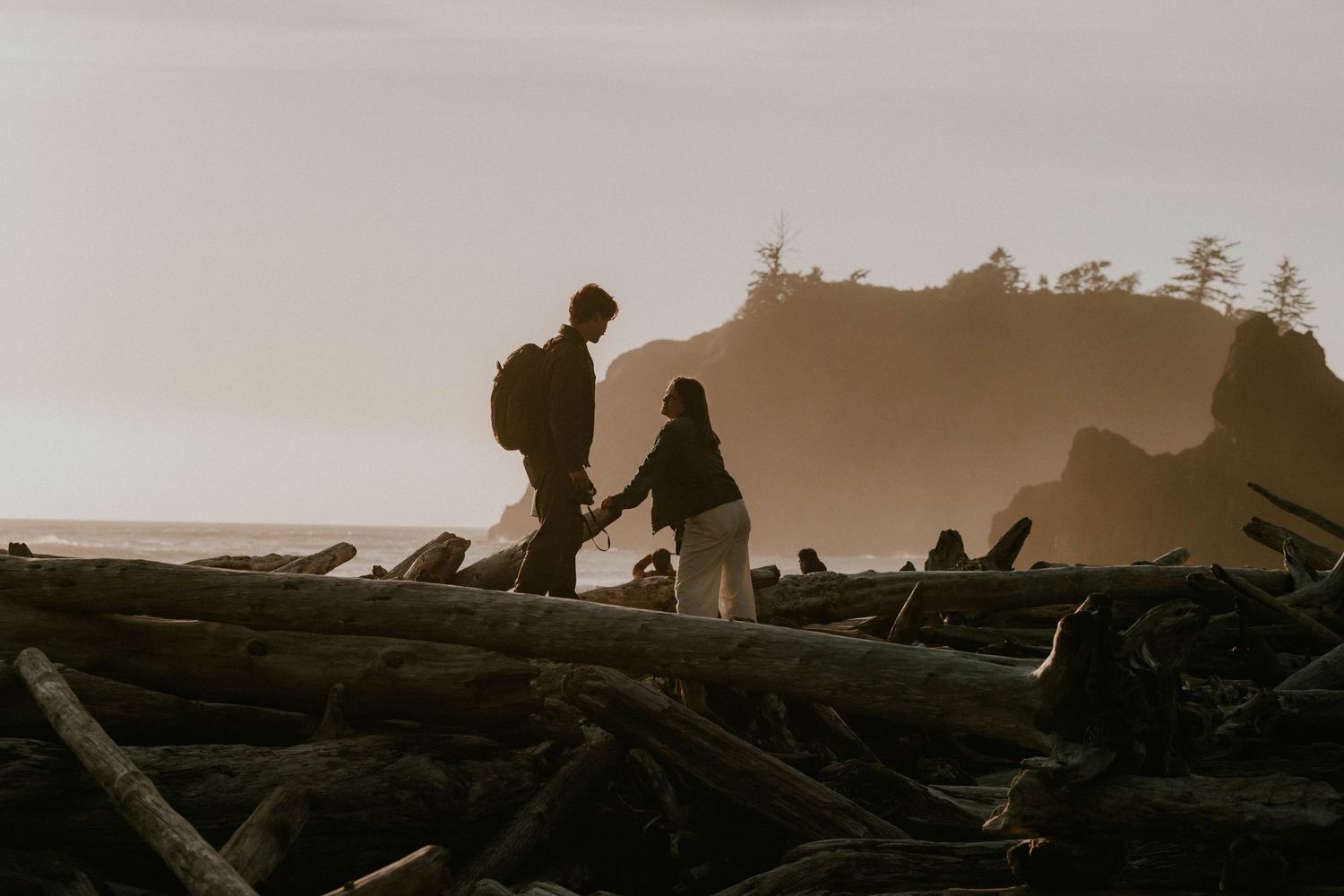 Couple holding hands while hiking over driftwood logs on a misty coastal beach at sunset with rocky cliffs in background.