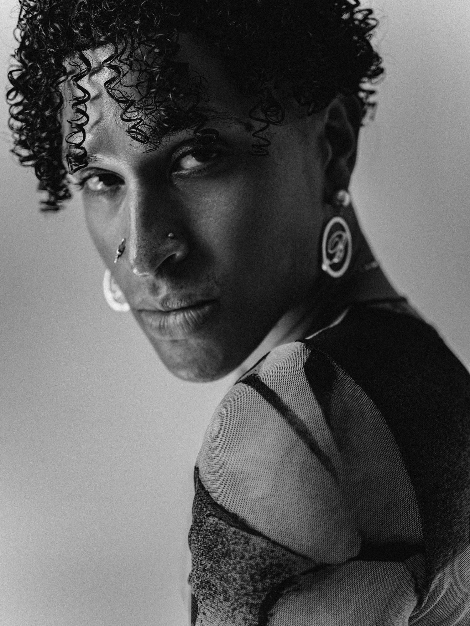 Black and white portrait of a person with curly hair, nose ring, and ear gauges looking over their shoulder.