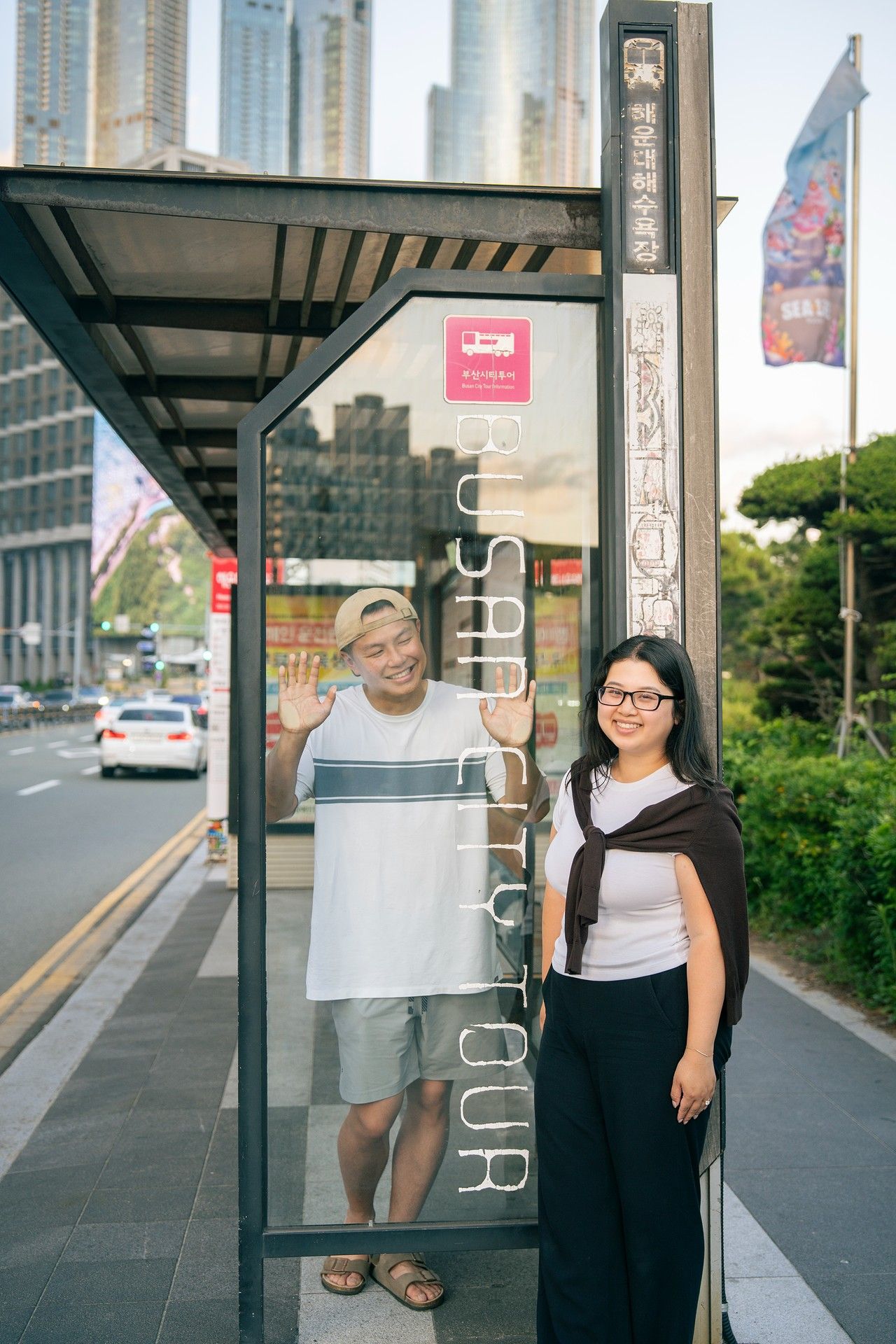 Two people waving and smiling at a Busan City Tour bus stop with urban skyline in background.